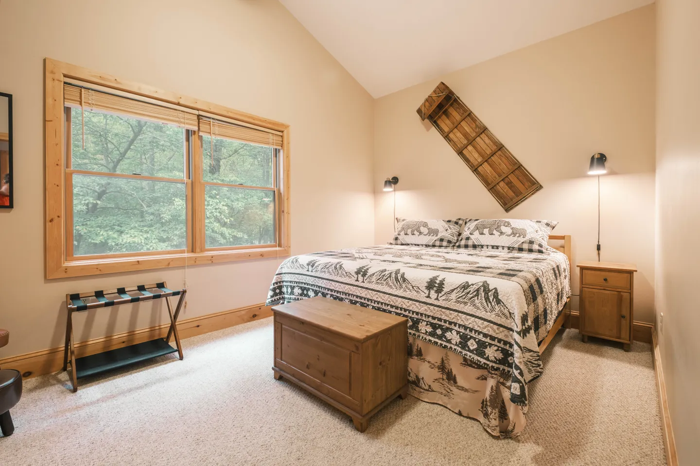 Bedroom with vaulted ceiling, carpeted floor, bed with patterned bedding, and large windows looking out to trees.