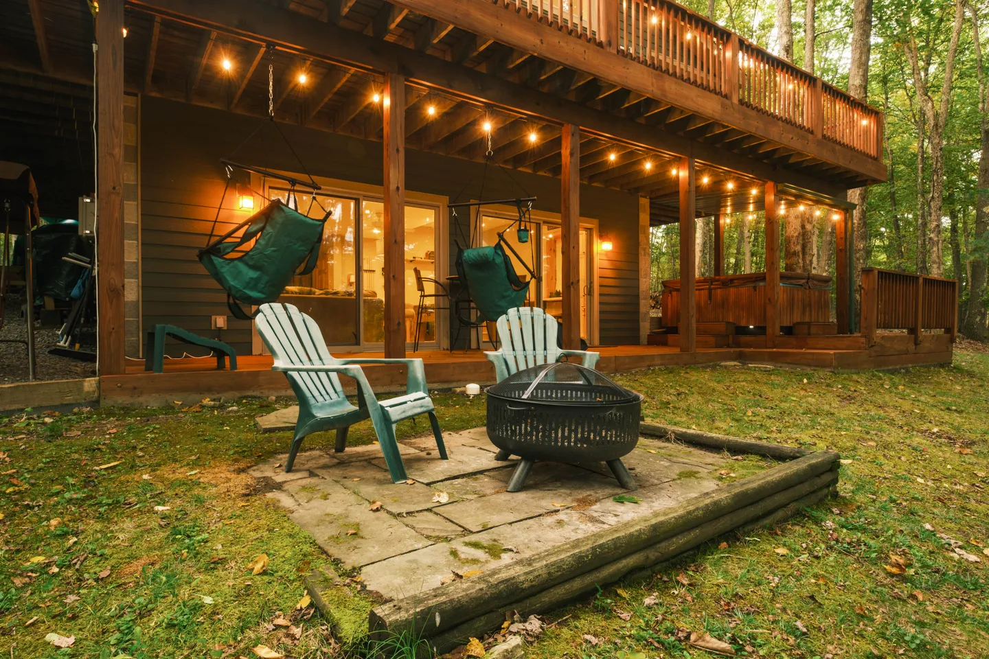 A backyard patio with a fire pit, Adirondack chairs, and string lights hanging from an elevated wooden deck above a lower porch area.