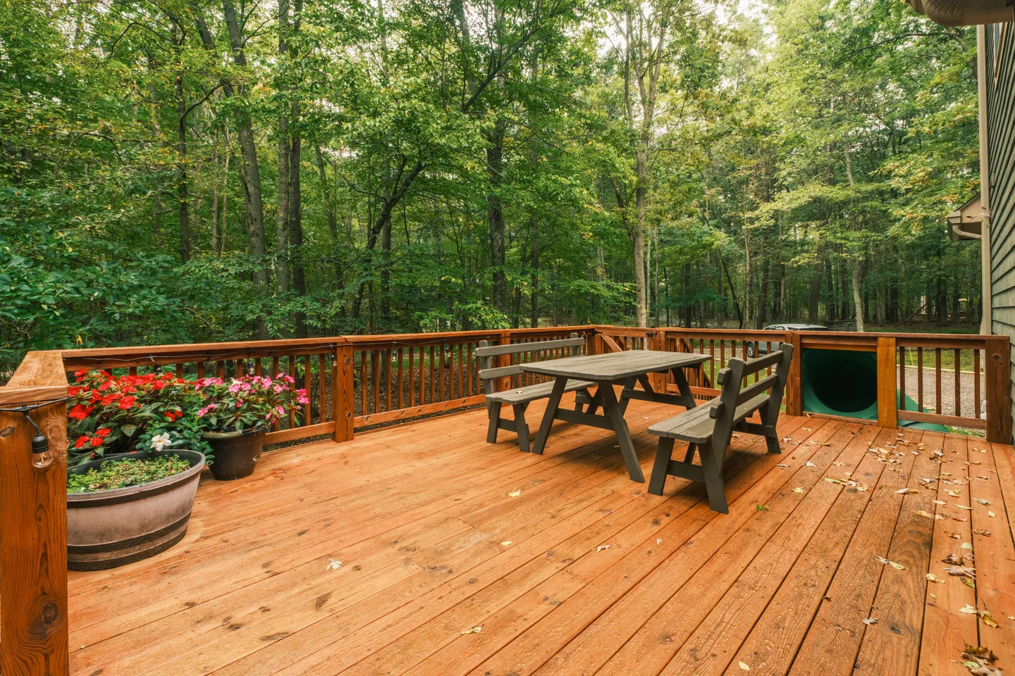 A wooden outdoor deck with a picnic table, potted flowers, and railings overlooking dense green trees.