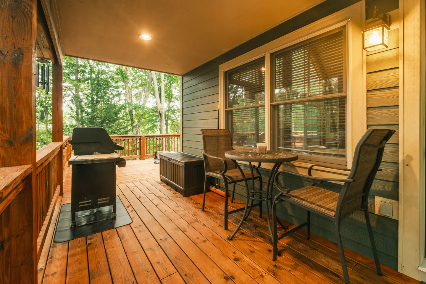 Covered wooden deck featuring a small table with two chairs, a barbecue grill, and a view of surrounding trees.