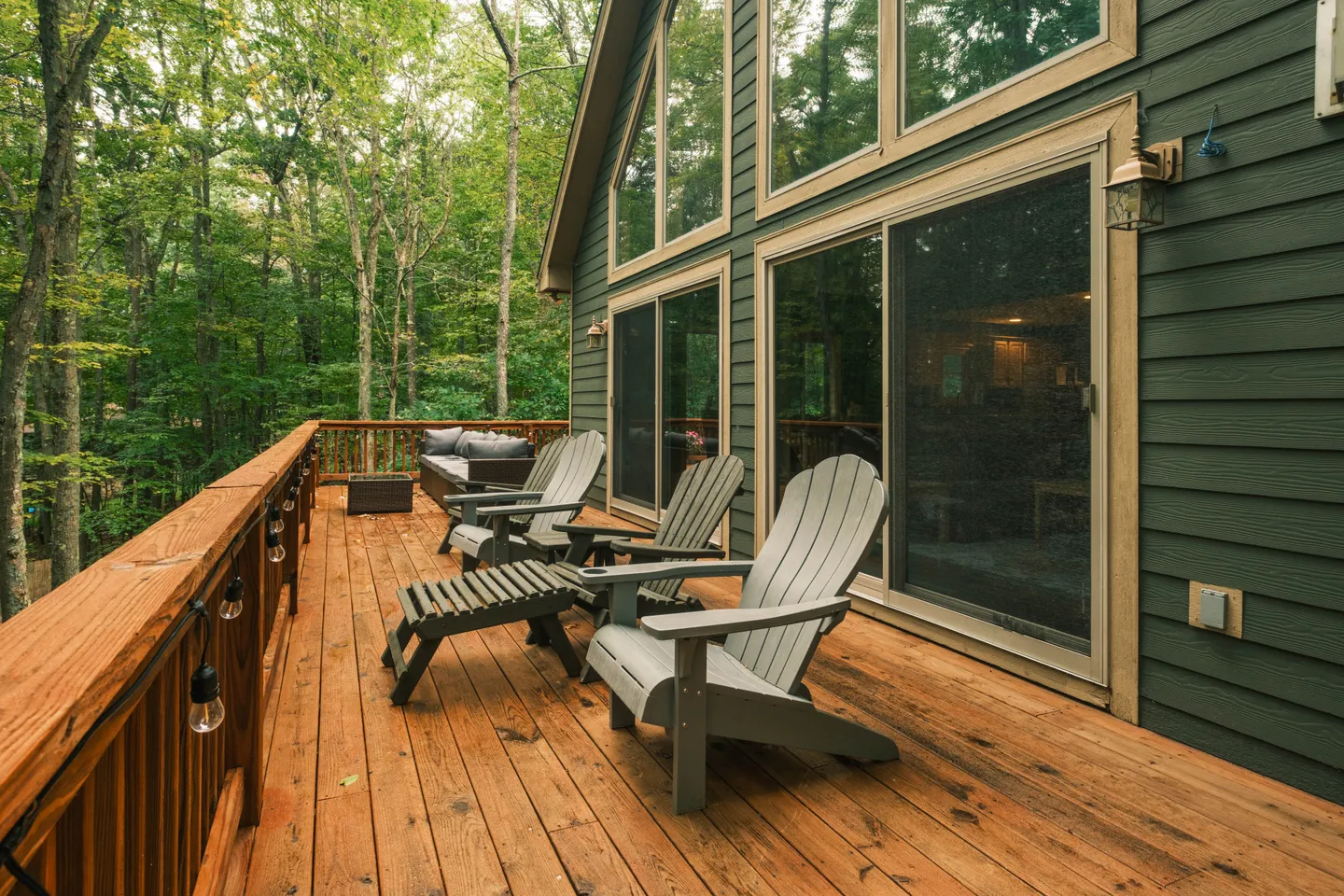 Wooden elevated deck with Adirondack chairs, an outdoor sofa, and string lights overlooking surrounding trees.