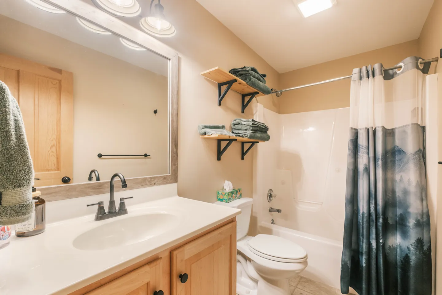 Bathroom with a wood-framed mirror over a single-sink vanity, a toilet, open wall shelves, and a shower-tub combo with a printed shower curtain.