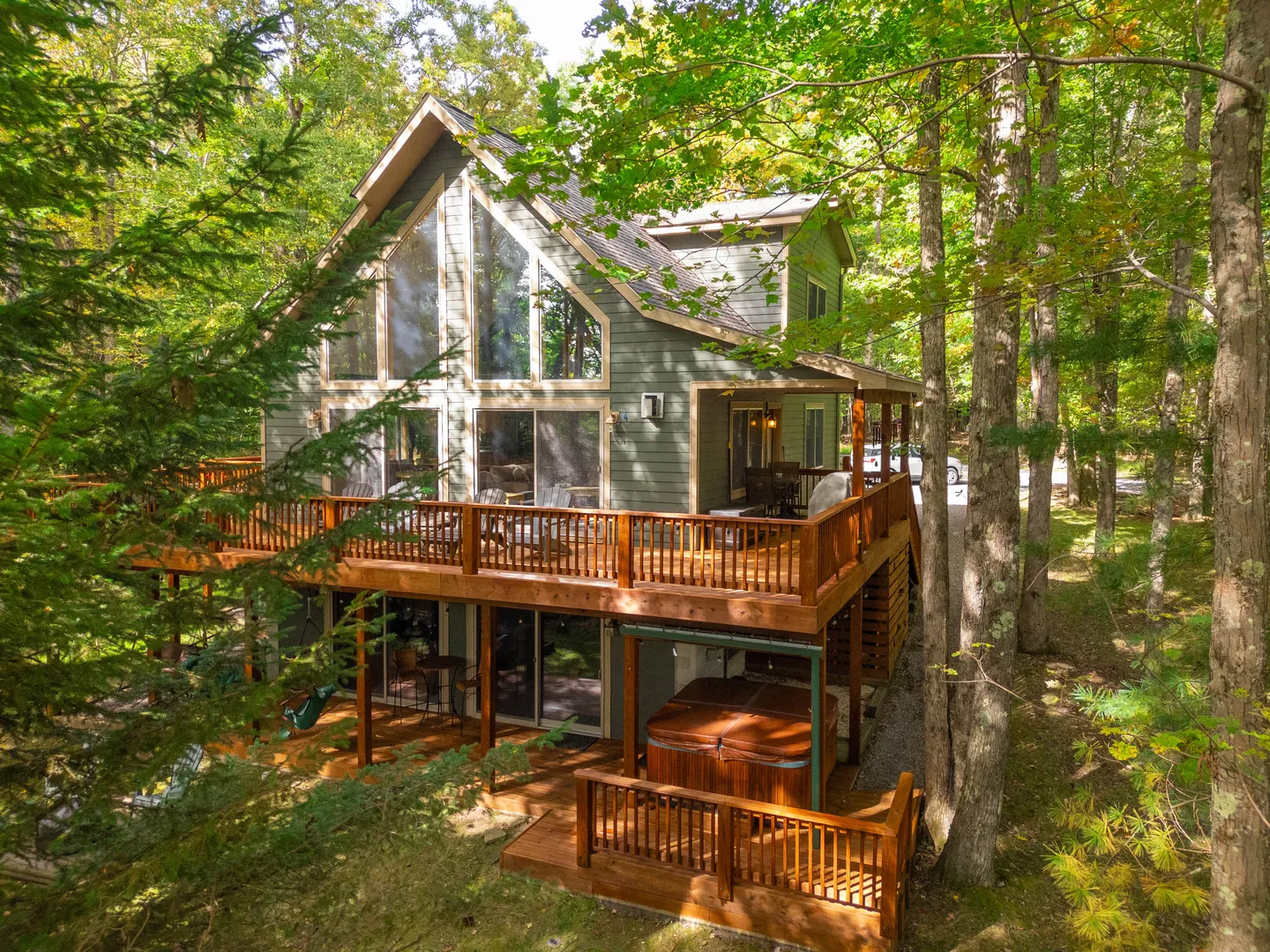 Exterior rear view of a two-story wooden cabin nestled in a forest, featuring multi-level wraparound decks, a hot tub, and large windows.