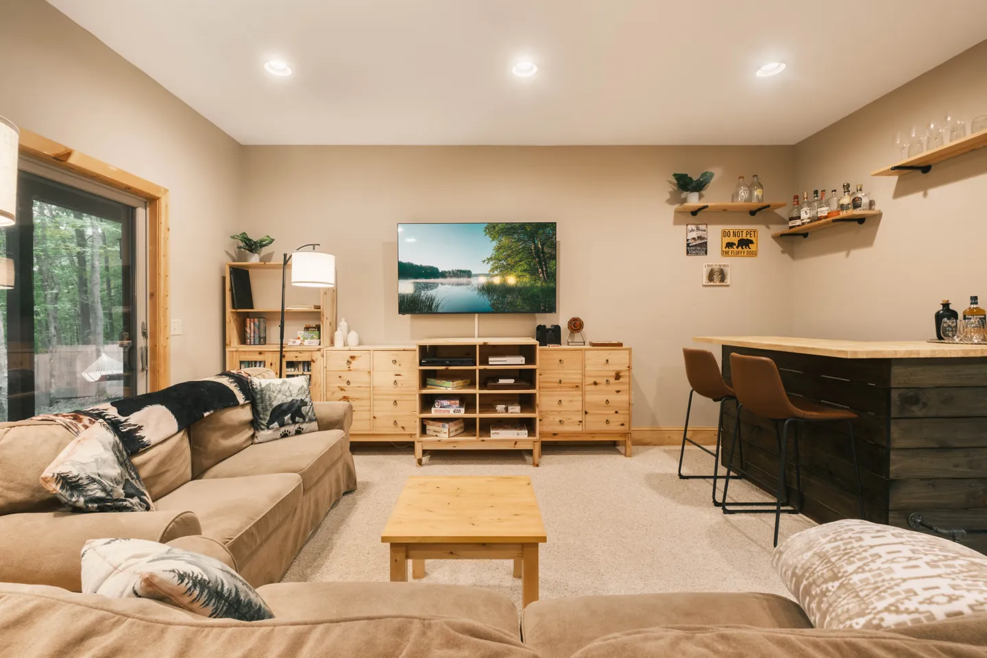 Living room and bar area with a tan sectional sofa, coffee table, light wood media console beneath a wall-mounted TV, and a wooden home bar with barstools.