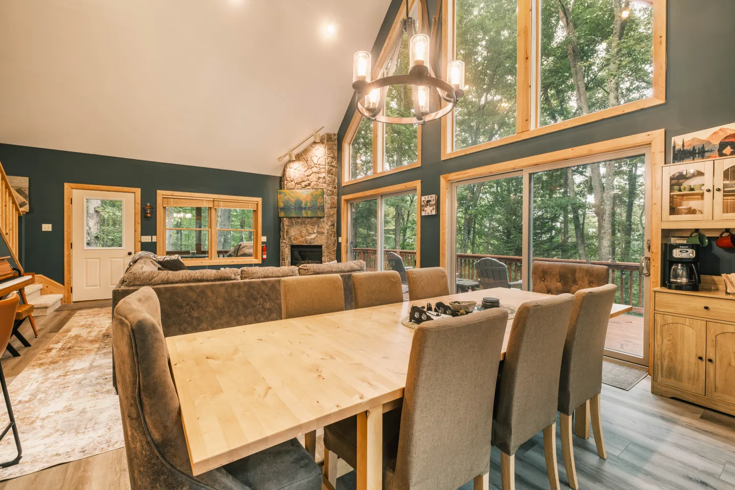 Open-concept dining and living room featuring a long wooden dining table, vaulted ceilings, and a tall wall of windows facing a wooded exterior deck.