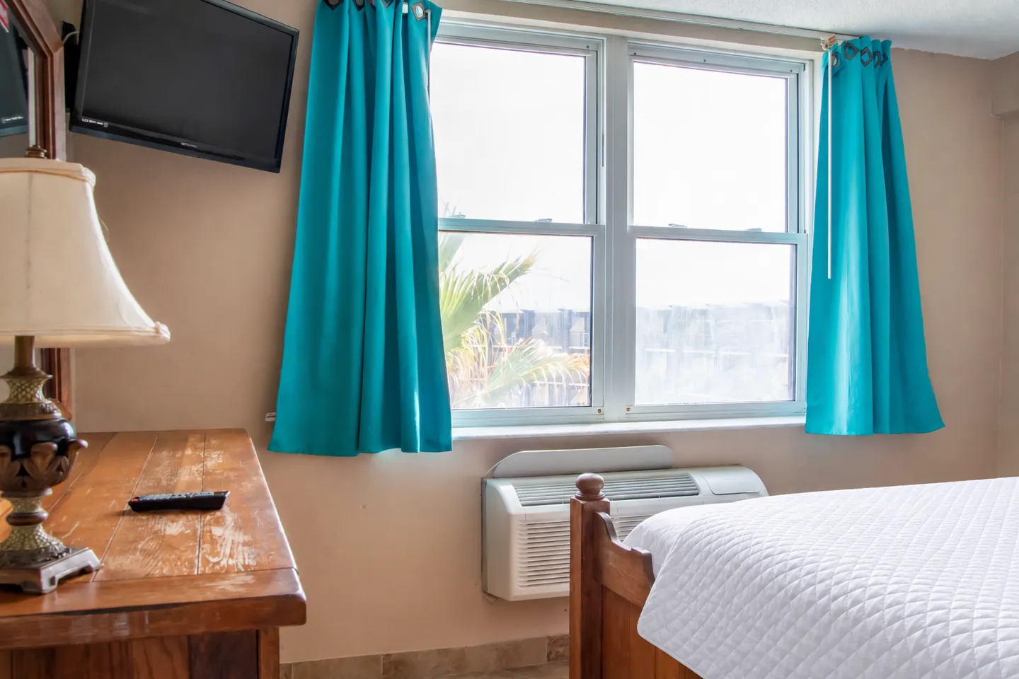 Bedroom featuring a window with bright blue curtains, an air conditioning unit beneath, a bed with a white quilt, a wooden dresser, and a wall-mounted TV.