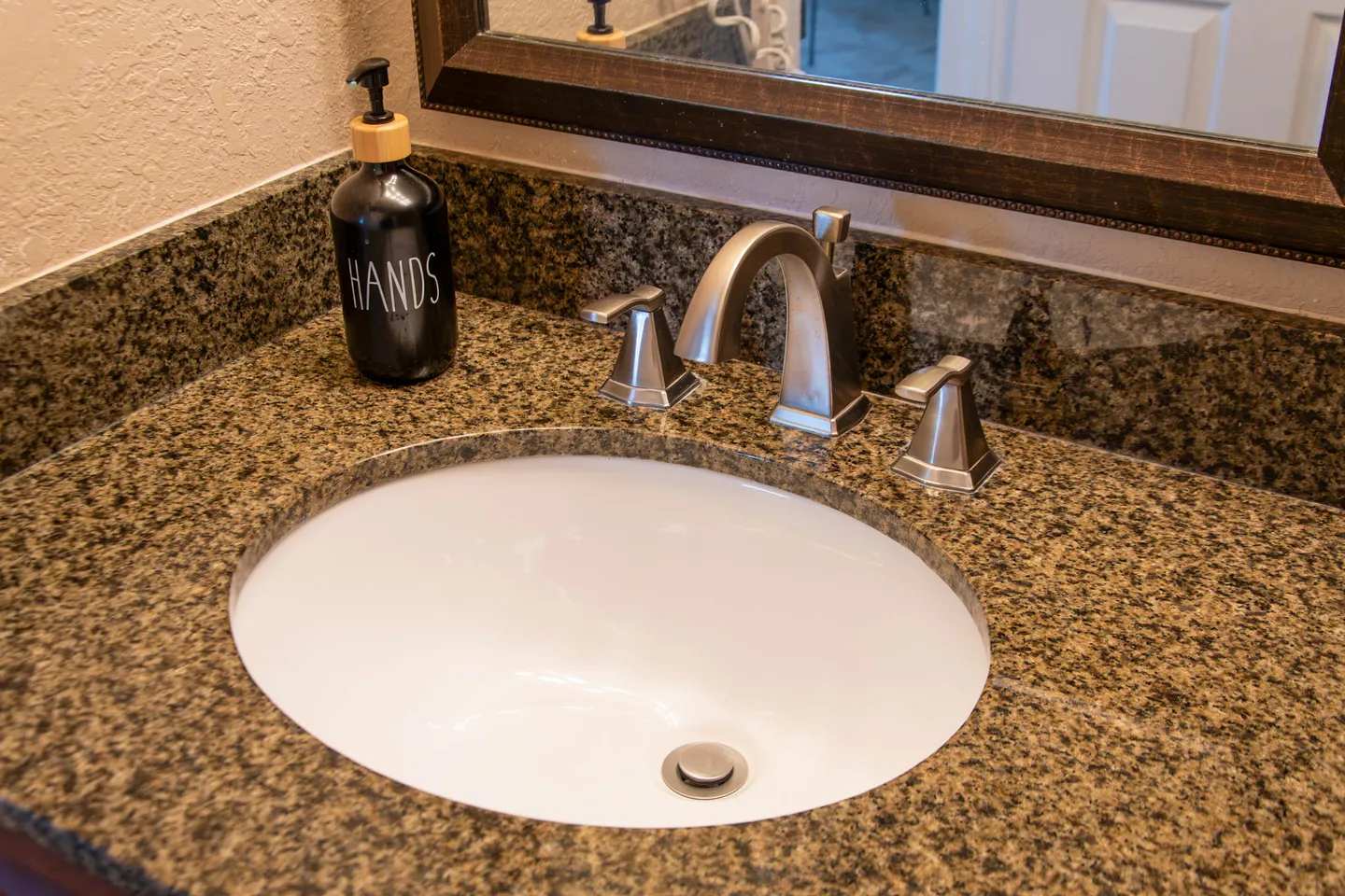 Close-up of a bathroom vanity with a speckled countertop, undermount oval sink, brushed metal faucet, and a black soap dispenser labeled HANDS.
