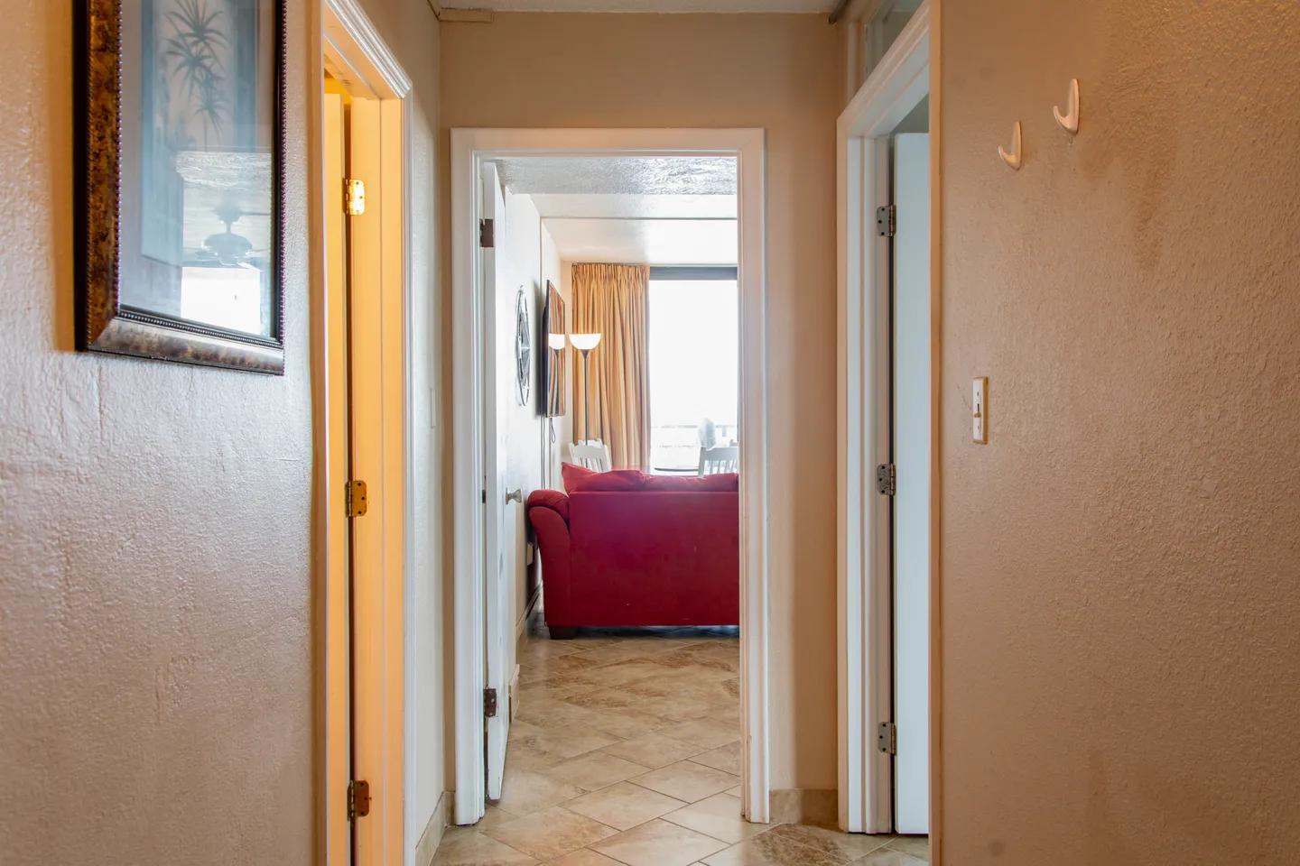 A tiled hallway looking past interior doorways toward a living area with a red sofa and bright window.
