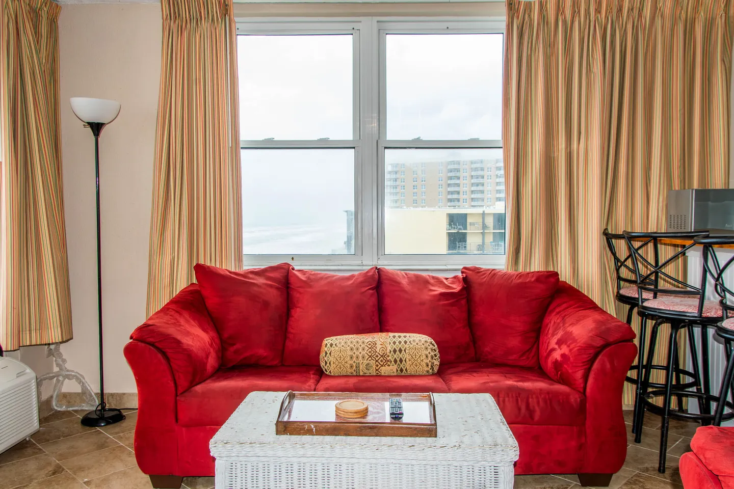 A bright red upholstered couch sits in front of a large window framed by striped curtains, with a white wicker coffee table in the foreground.