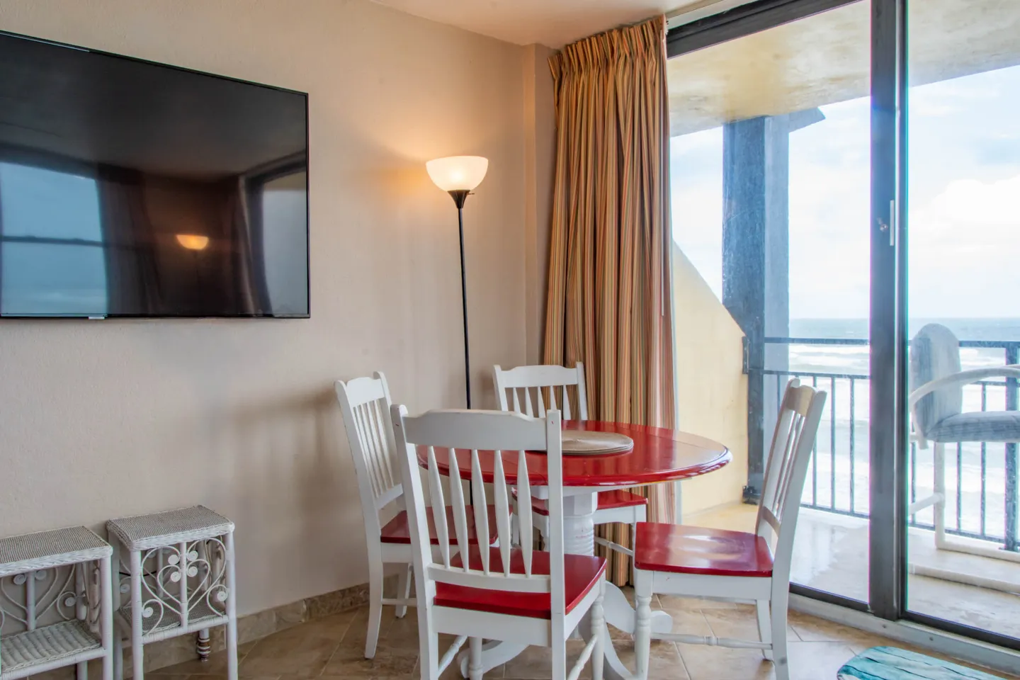 Dining area with a round red and white table and chairs next to a sliding glass door leading to a balcony with ocean views.