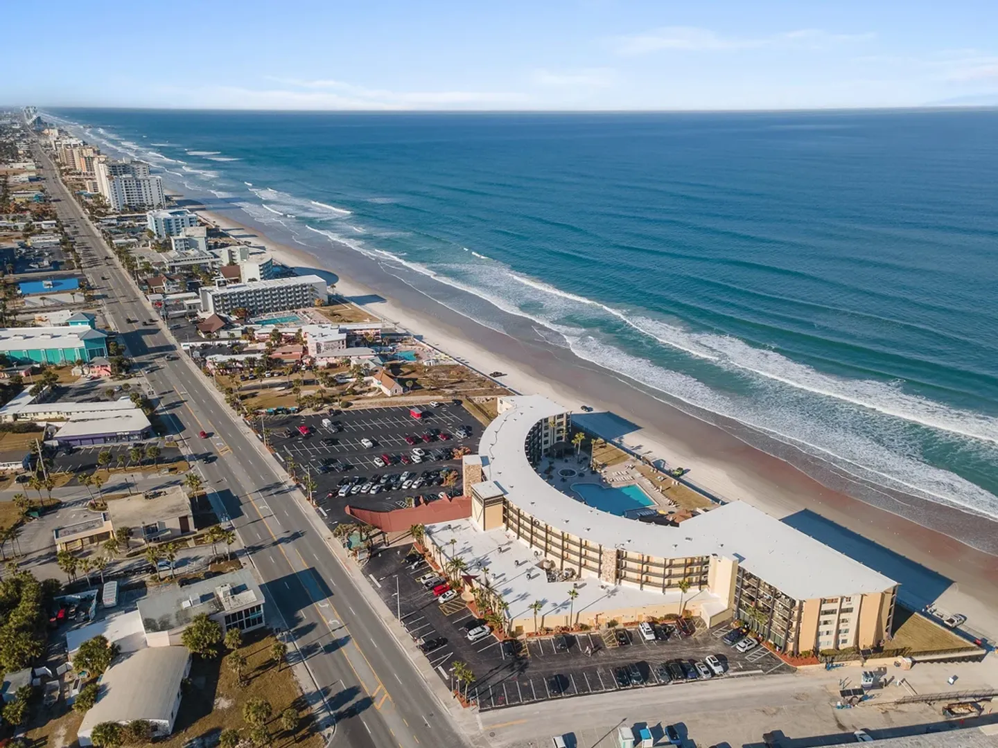 Aerial view of a curved oceanfront resort building directly bordering a sandy beach and ocean, flanked by a coastal road and parking lots.