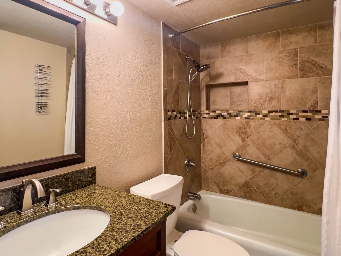 Bathroom with granite vanity countertop, framed mirror, toilet, and bathtub with tiled surround and shower head.