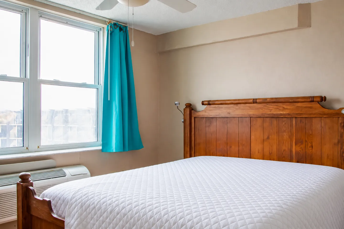 Bedroom with a wooden bed dressed in a white quilted coverlet next to a window with a turquoise curtain.