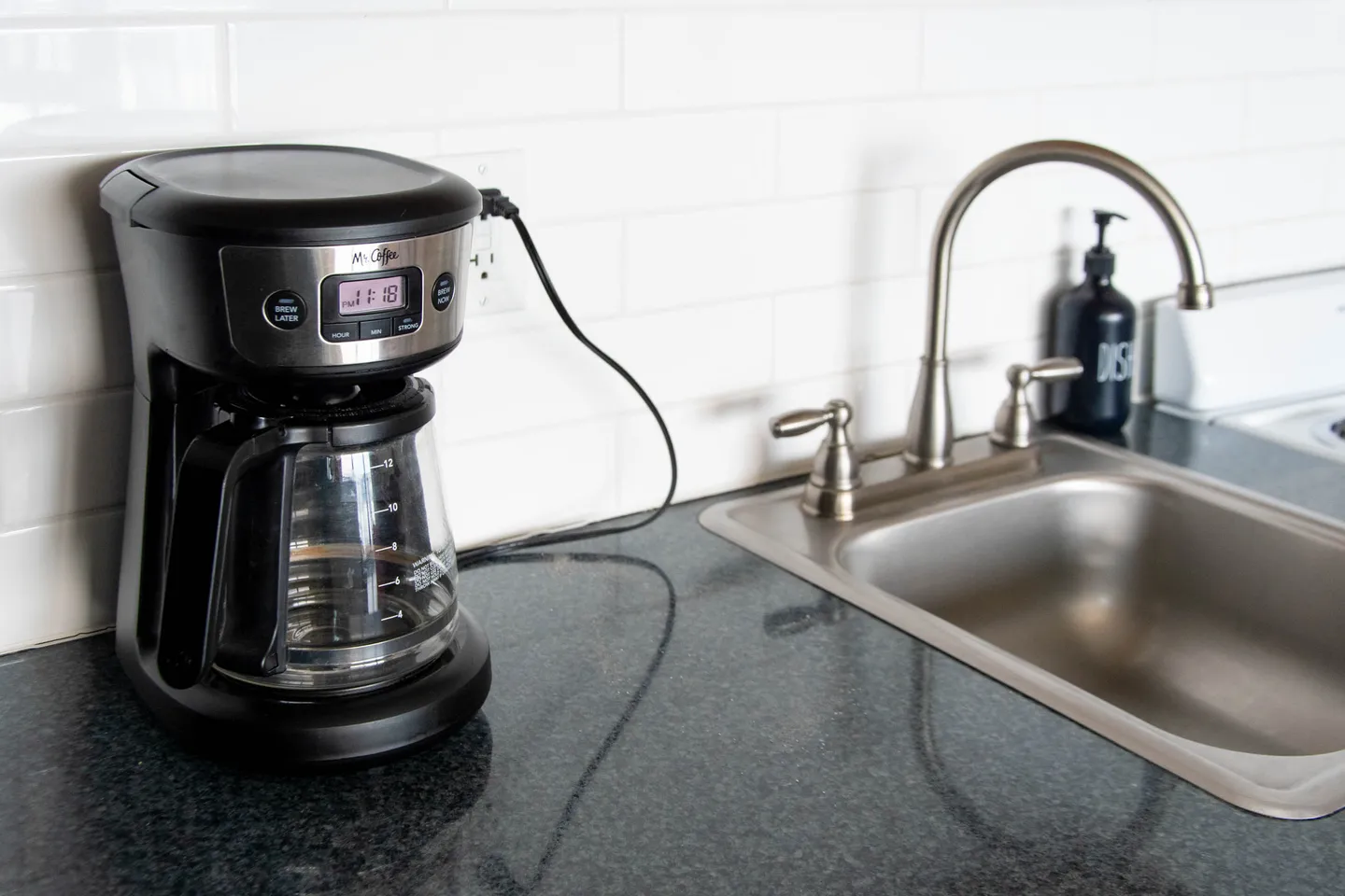 A black and stainless steel drip coffee maker plugged into the wall on a dark countertop next to a kitchen sink.