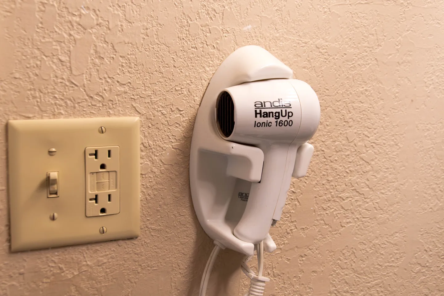 A wall-mounted white hair dryer in a cradle next to an electrical outlet with a light switch.