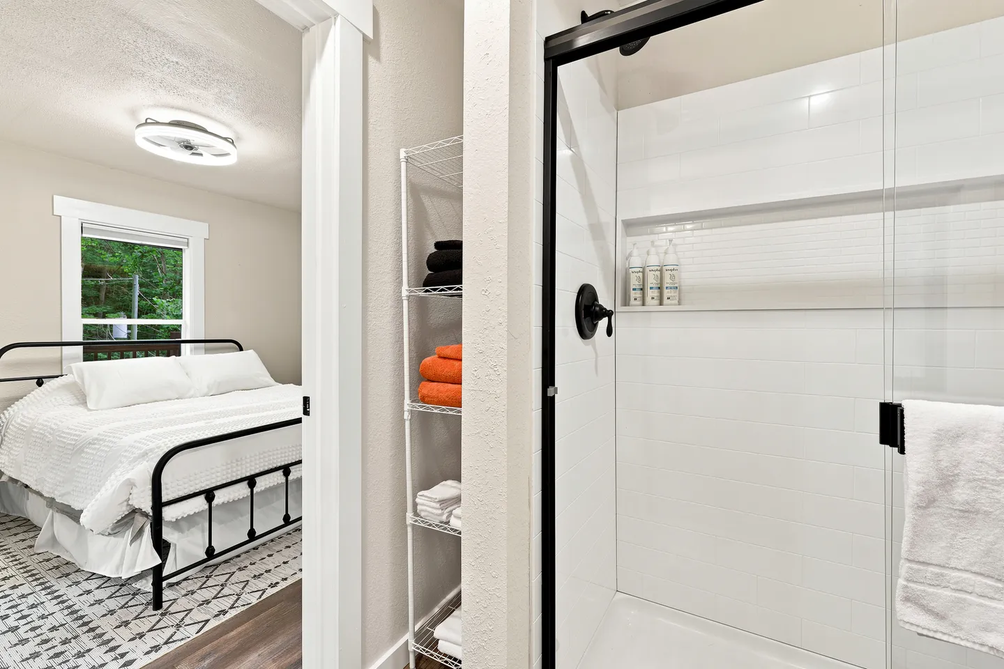 A view from a bathroom showing a walk-in shower with white tile and black trim, adjacent to an open doorway into a bedroom with a black metal bed.