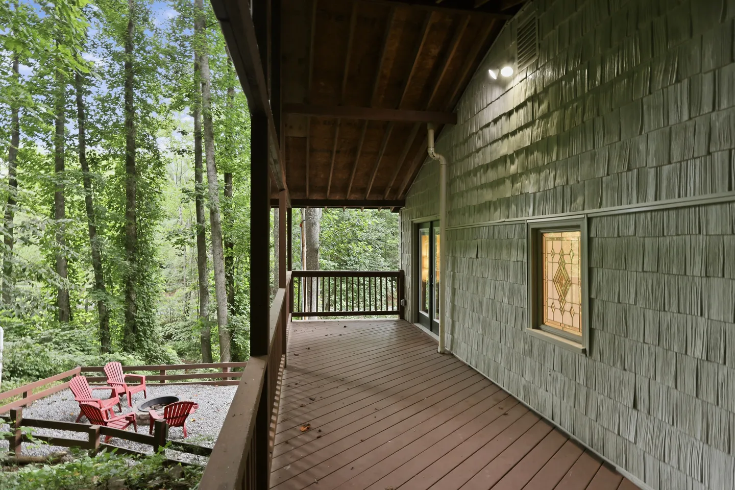 A covered wooden deck running alongside a home with shingle siding, looking out toward dense woods and a ground-level fire pit patio.