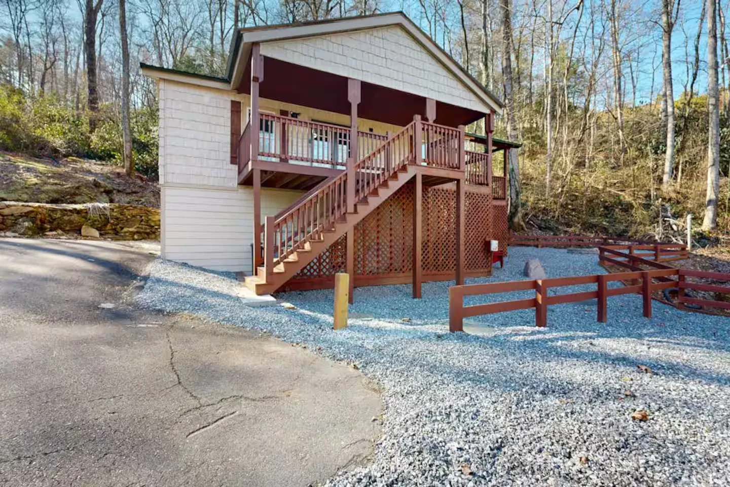 Exterior view of a multi-level wood-sided cabin with an elevated covered deck, exterior wooden stairs, and a gravel driveway area.