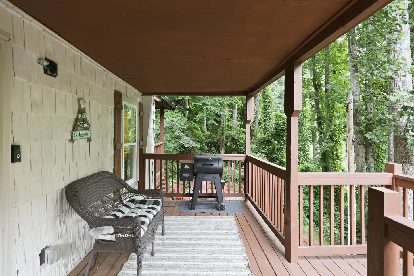 Covered wooden porch with a wicker bench, area rug, and outdoor grill overlooking trees.
