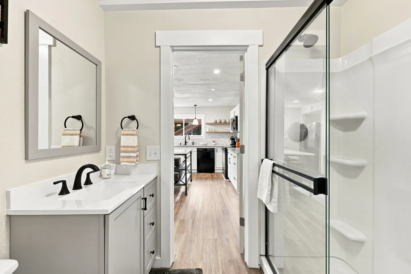 Bathroom with a grey vanity, framed mirror, and glass-door shower stall looking out an open doorway into the kitchen.