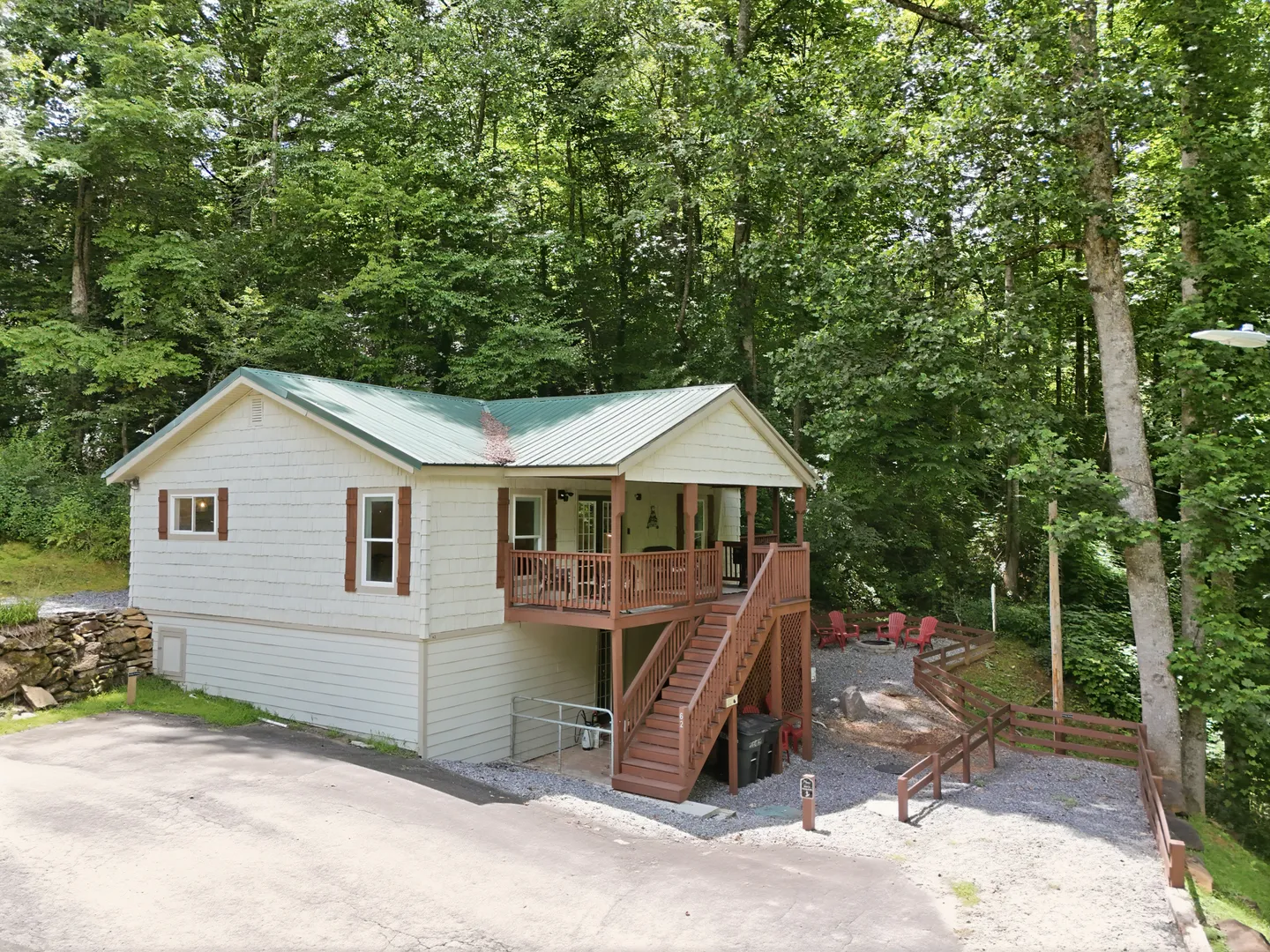 Exterior view of a light-colored cabin with a green metal roof, elevated wooden deck, and gravel yard surrounded by dense trees.