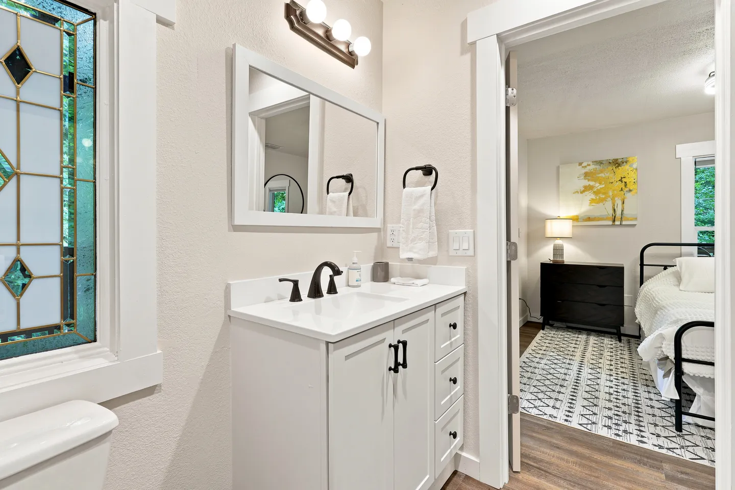 Bathroom with white vanity, mirror, stained glass window, and doorway opening to an adjoining bedroom.