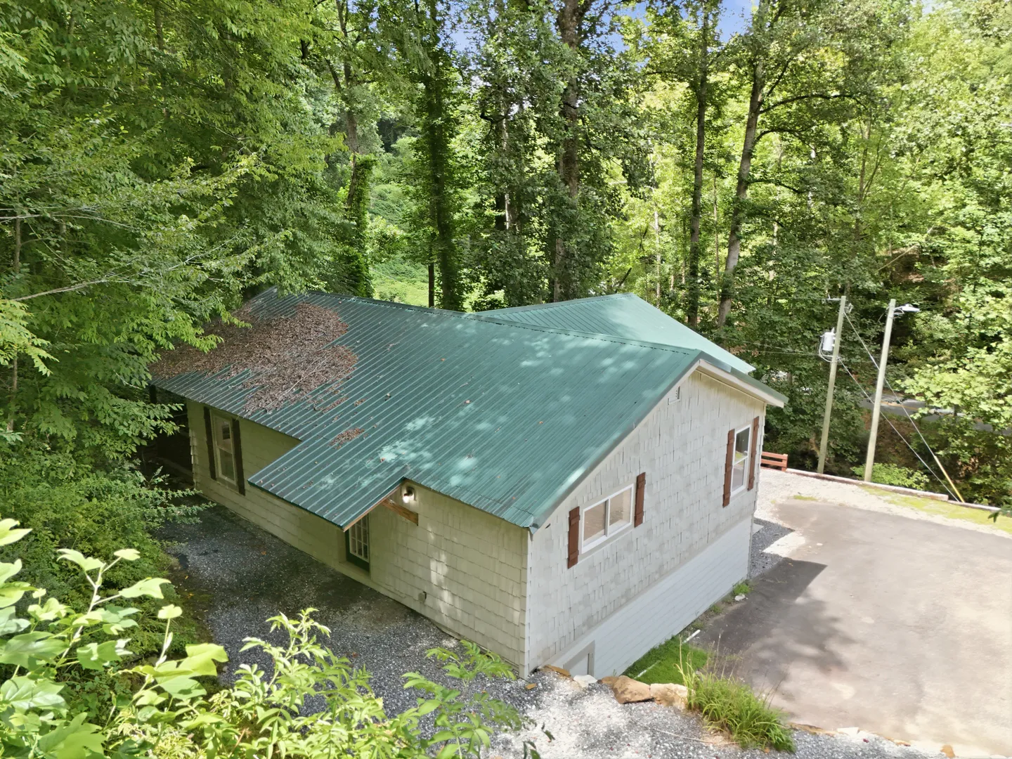 High-angle view of a house with a green metal roof surrounded by tall green trees and gravel driveway.