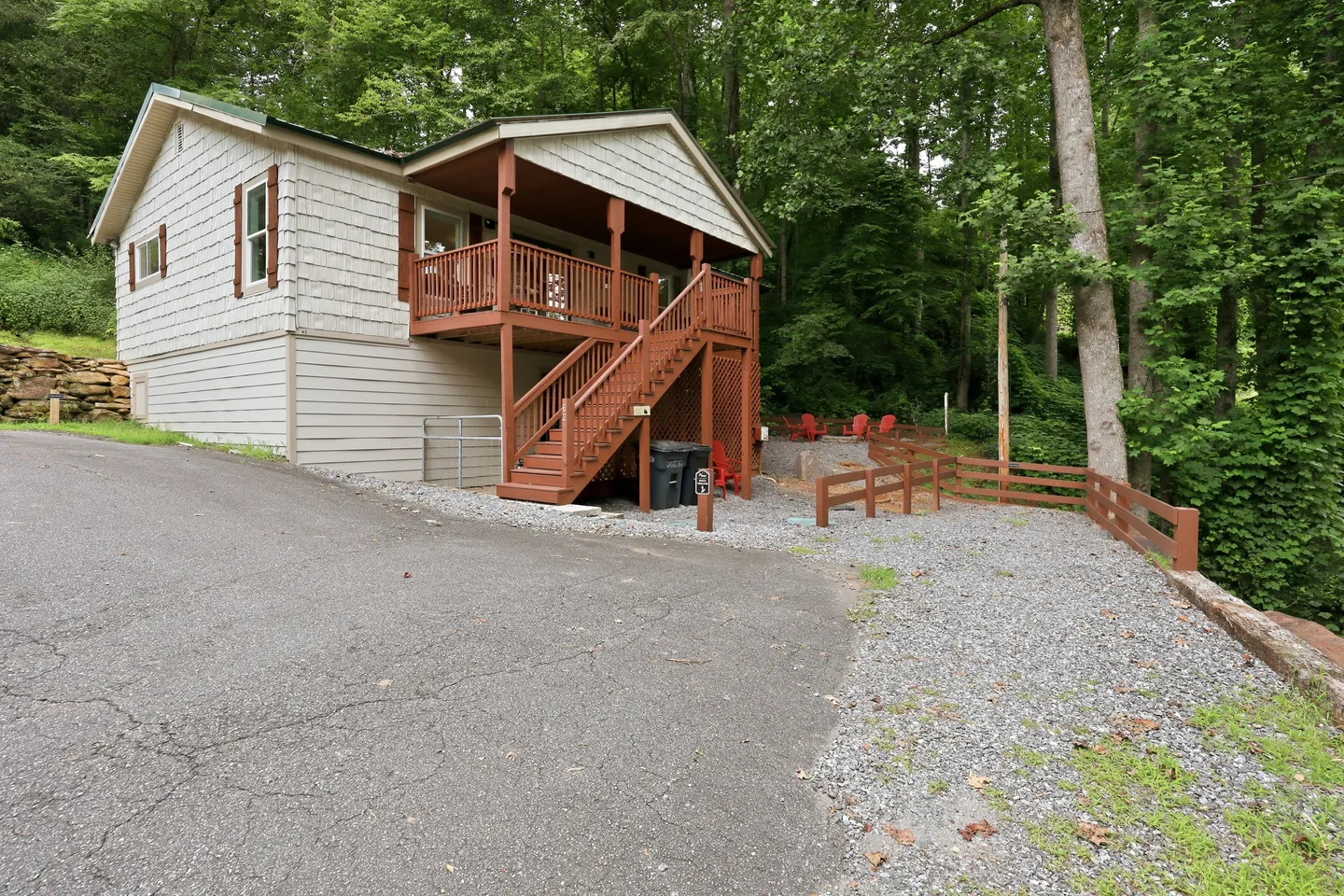 Exterior view of a cabin with light siding, a raised wooden porch with stairs, and a paved and gravel driveway surrounded by trees.
