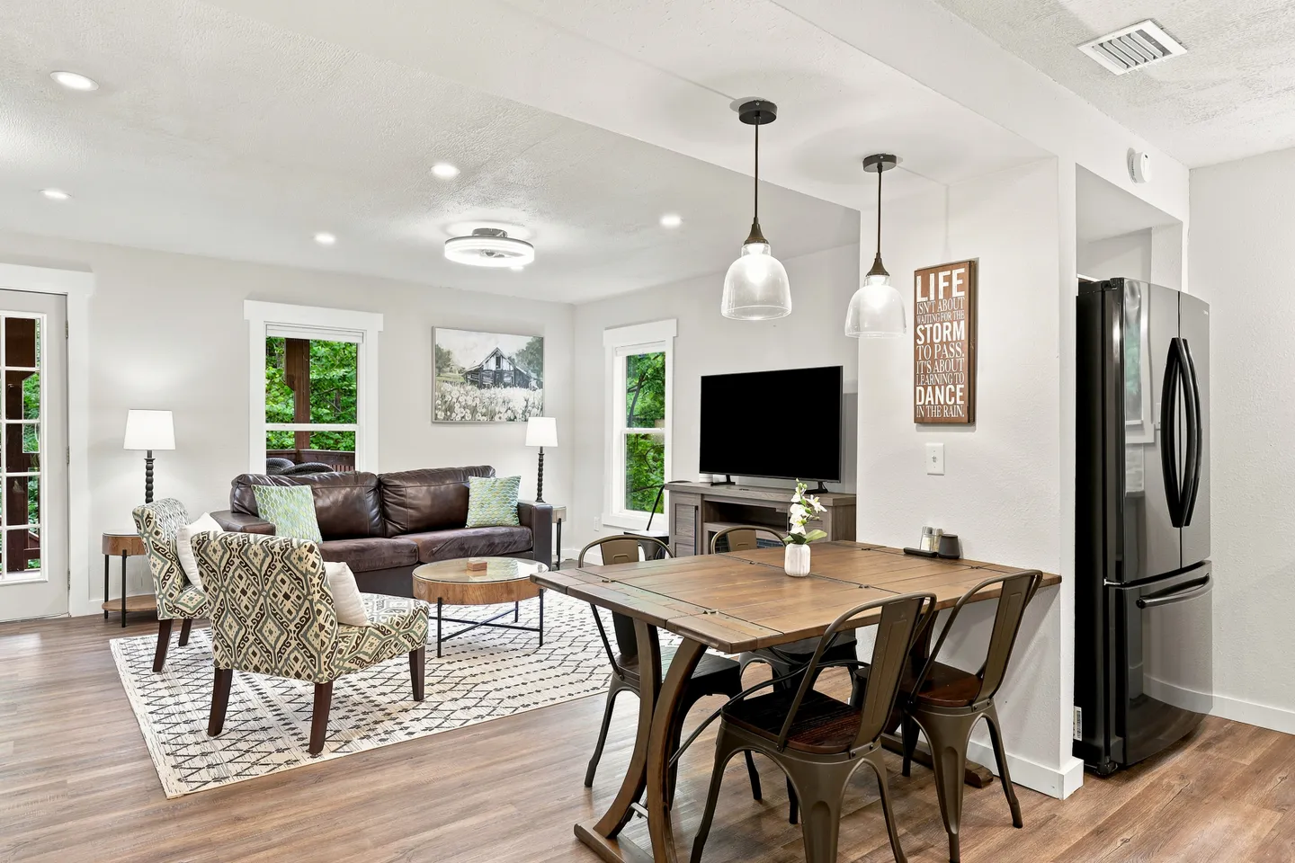 Open-concept living and dining area featuring wood-style flooring, a wooden dining table with metal chairs, a leather sofa, patterned armchairs, and a wall-mounted TV.