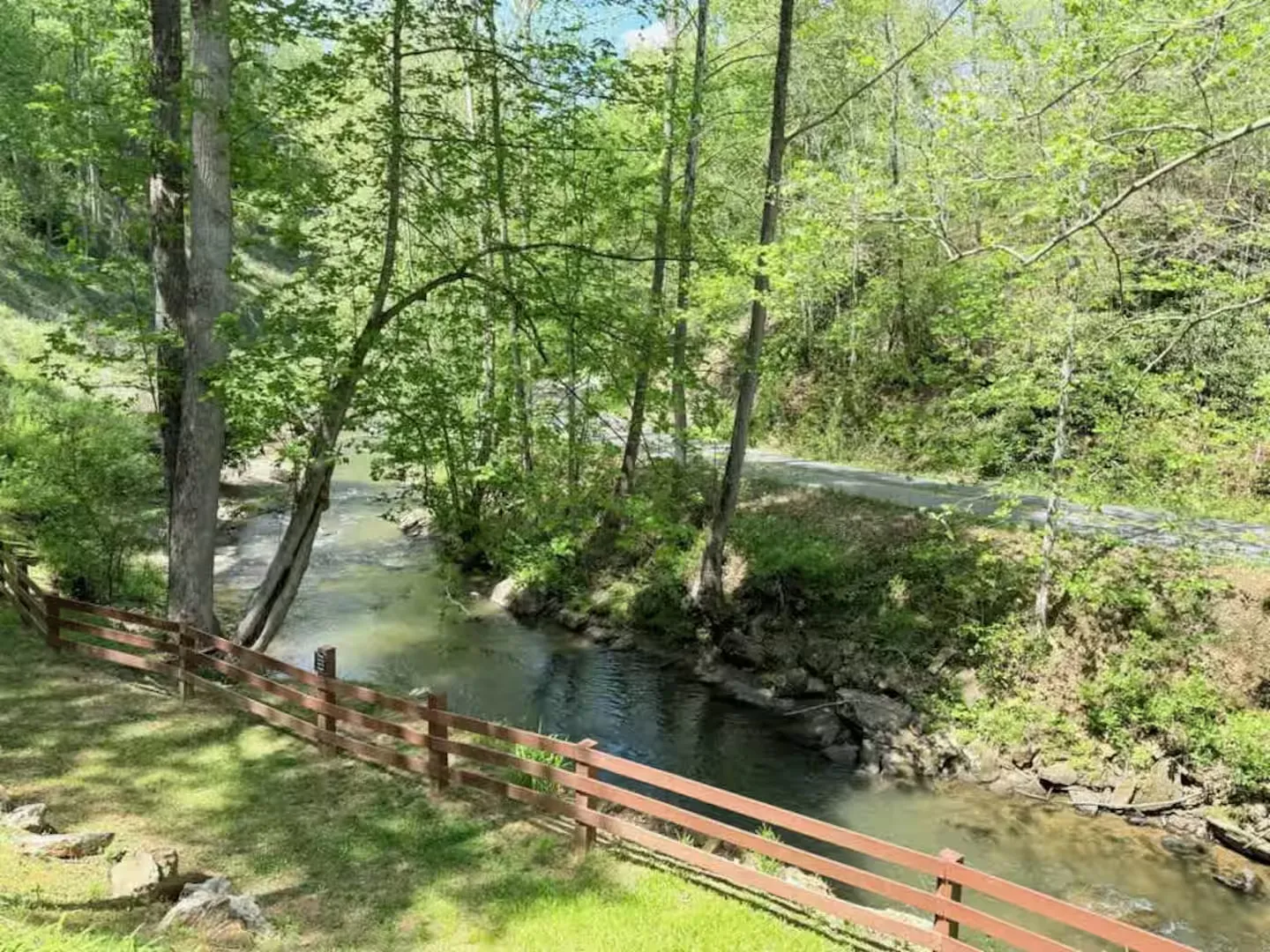 A flowing shallow stream bordered by a wooden fence, grassy slope, and lush green trees.