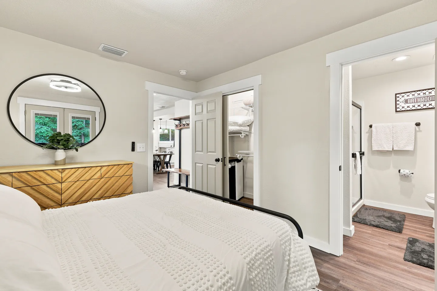 Bedroom with a bed made with white textured bedding, a wooden dresser with a circular mirror, and doorways to a hallway, closet, and en-suite bathroom.