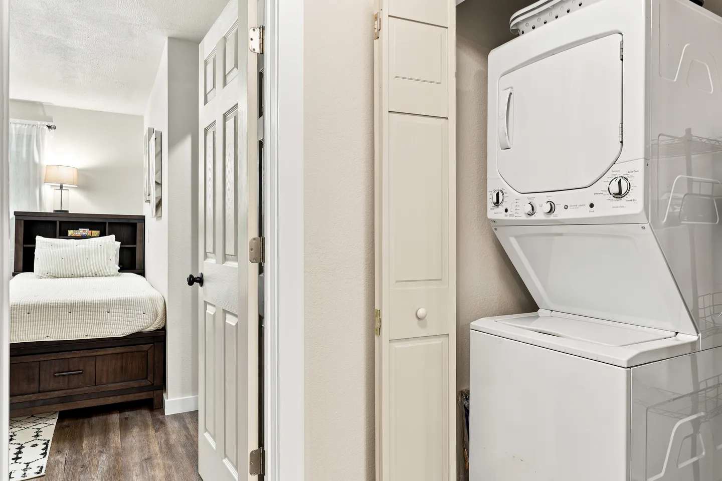 A stacked white washer and dryer in a hallway closet with a doorway revealing a bedroom beyond.