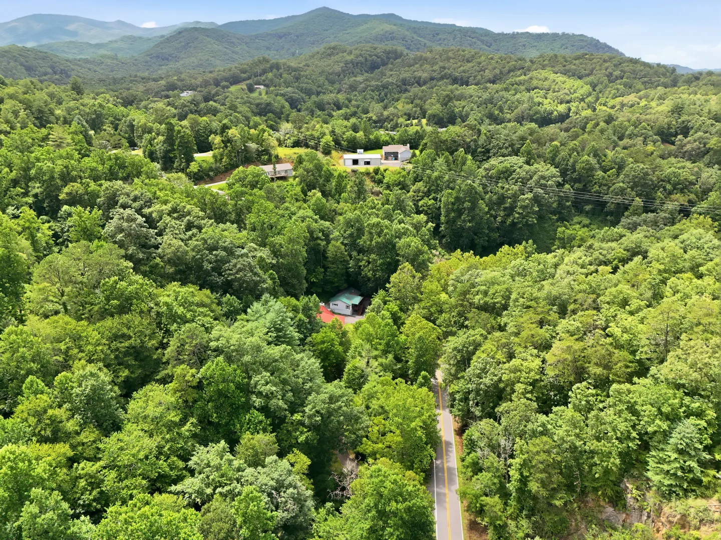 Aerial view of a paved road cutting through dense green forest leading toward cabins nestled in the hills.