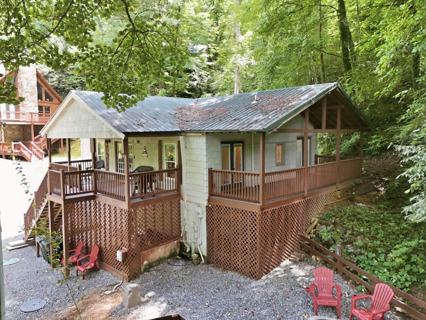 Exterior view of an elevated cabin surrounded by dense green trees with a wraparound wooden deck and lattice skirting.