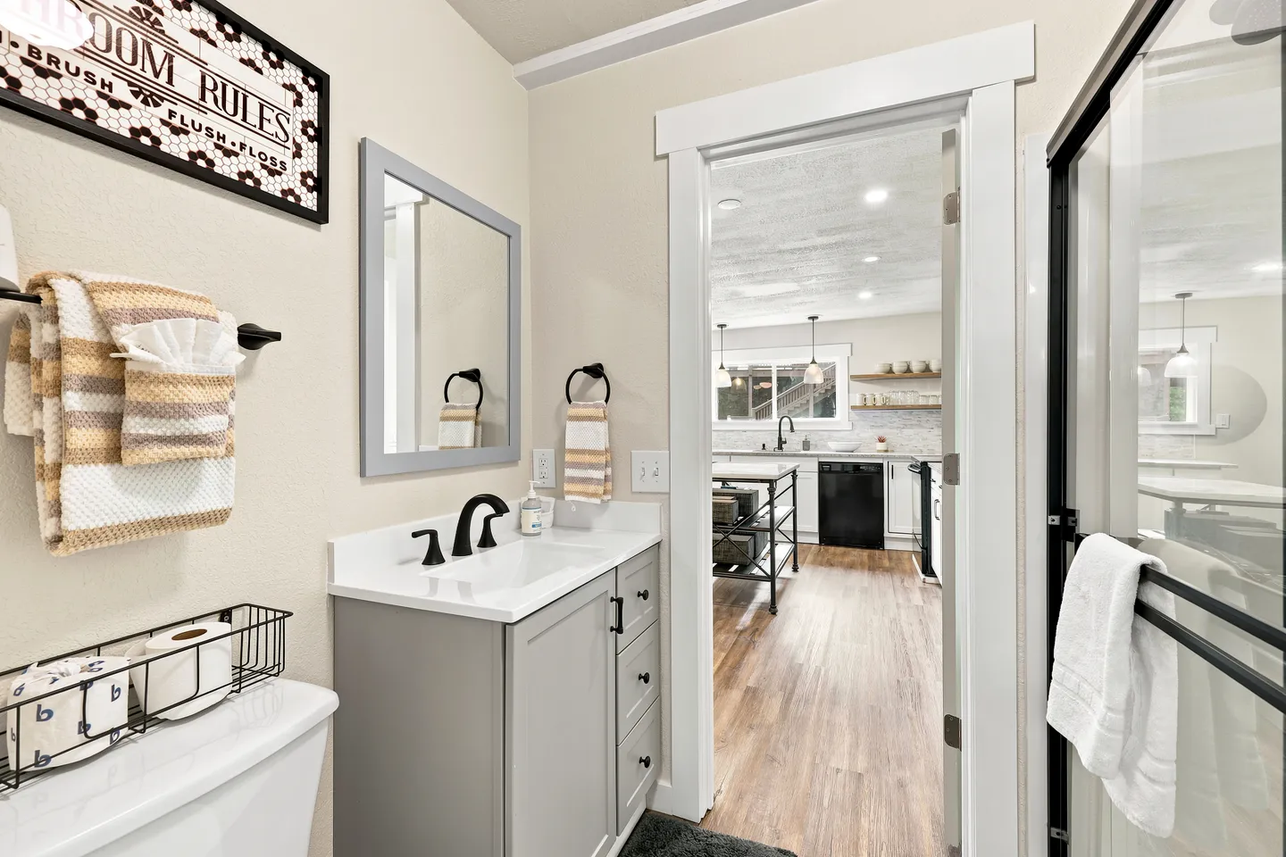Bathroom with a grey vanity, white countertop, framed mirror, and an open doorway looking out toward the kitchen.