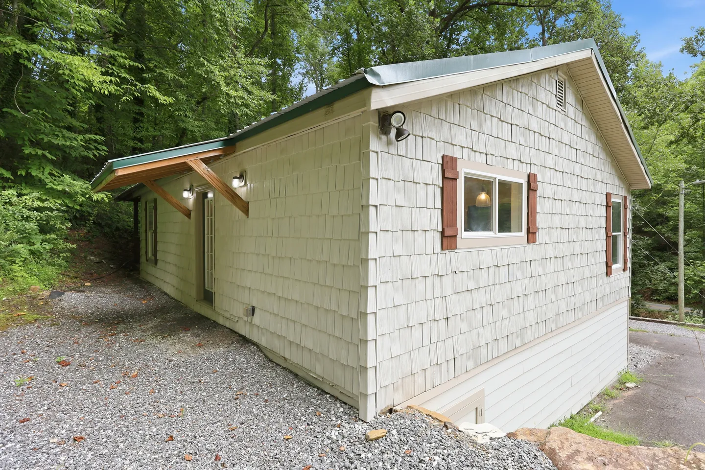 Exterior view of a cabin-style building with light-colored shake siding surrounded by trees and a gravel driveway.