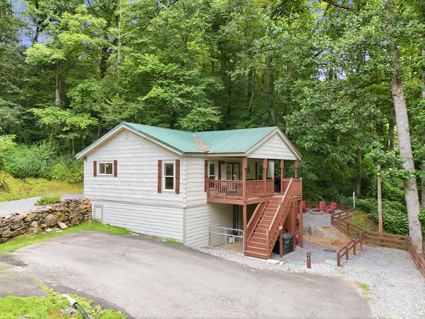 Exterior view of a hillside home with light-colored siding, a green metal roof, a wooden elevated porch with stairs, and a paved driveway surrounded by woods.
