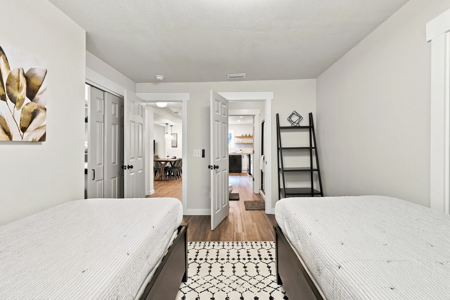 Bedroom with two beds, wood-look flooring, a patterned rug, and open doors leading to the hallway and bathroom.