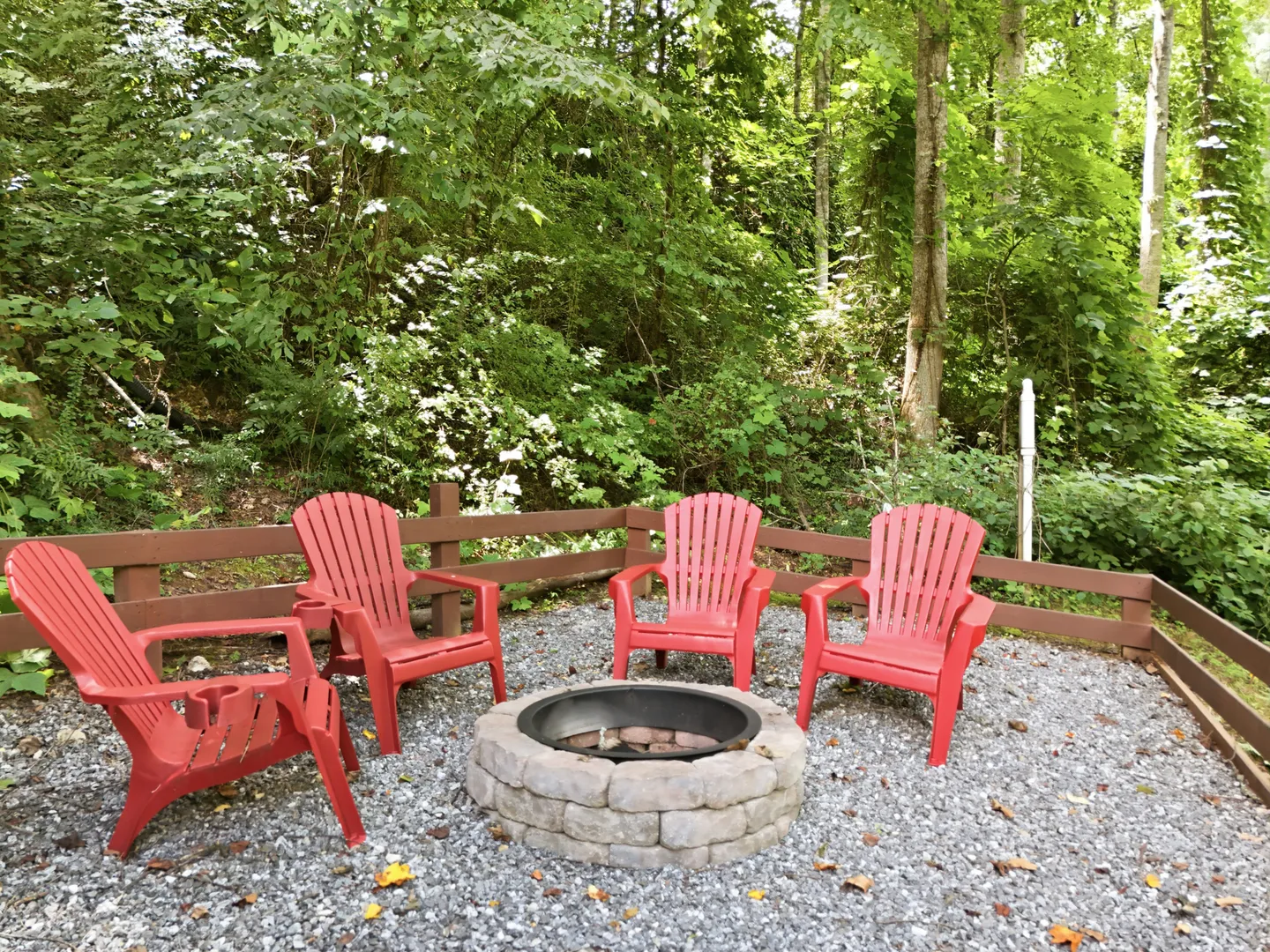 Outdoor gravel fire pit area with red Adirondack chairs bordered by a wooden fence and lush green woods.