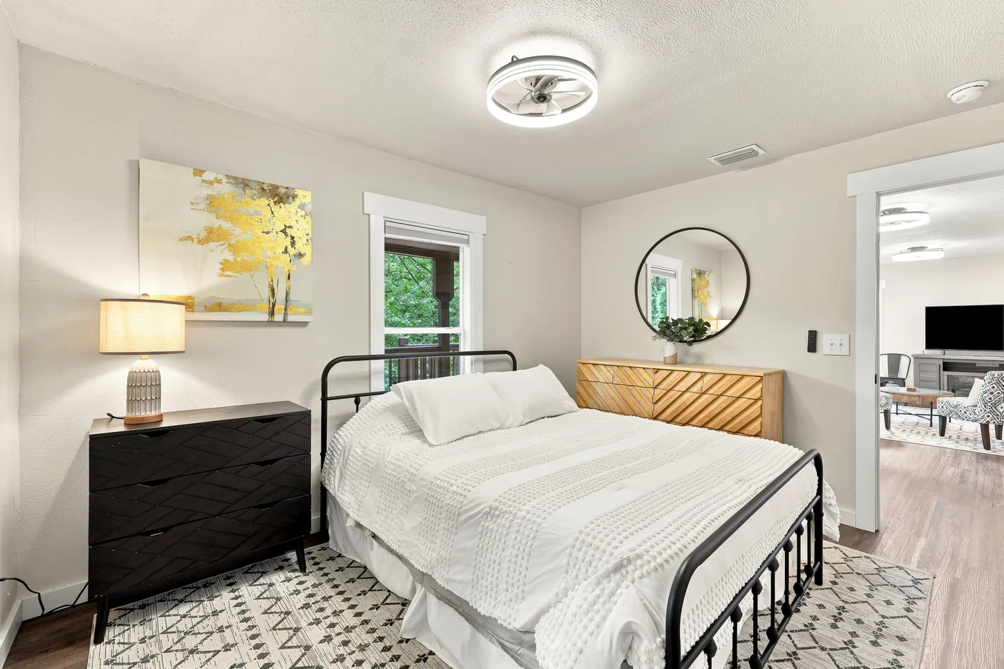 Bedroom with a black metal bed frame, patterned rug, dressers, and a doorway showing a living area.