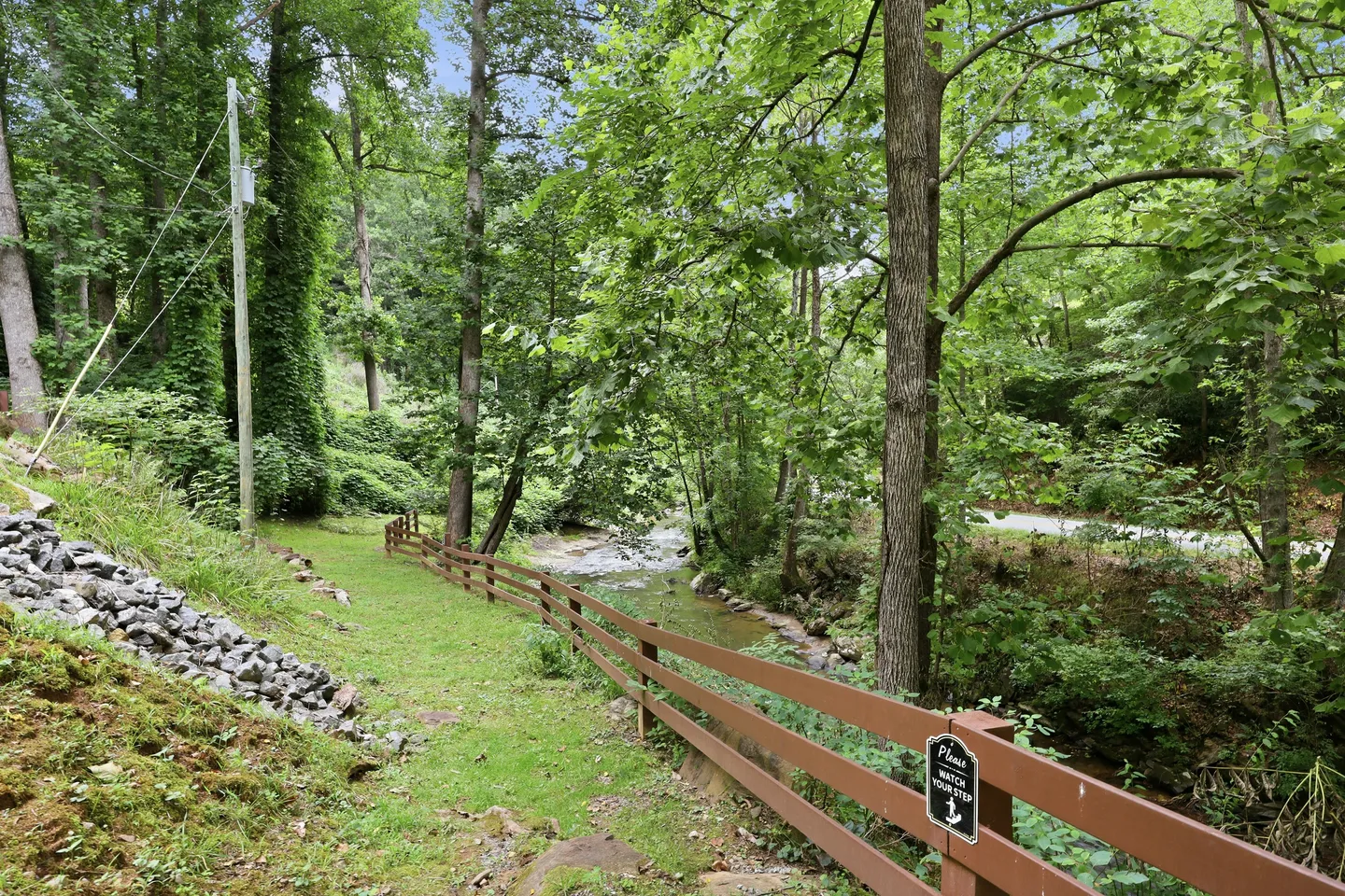 A brown wooden fence borders a grassy path running alongside a shallow stream surrounded by dense green trees.