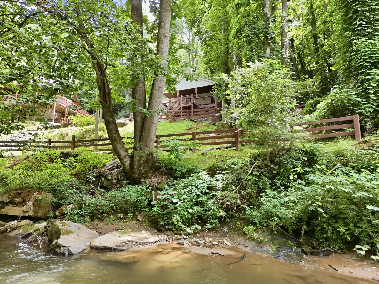A wooded hillside with a shallow stream in the foreground leading up toward a cabin with a porch and wooden fencing.
