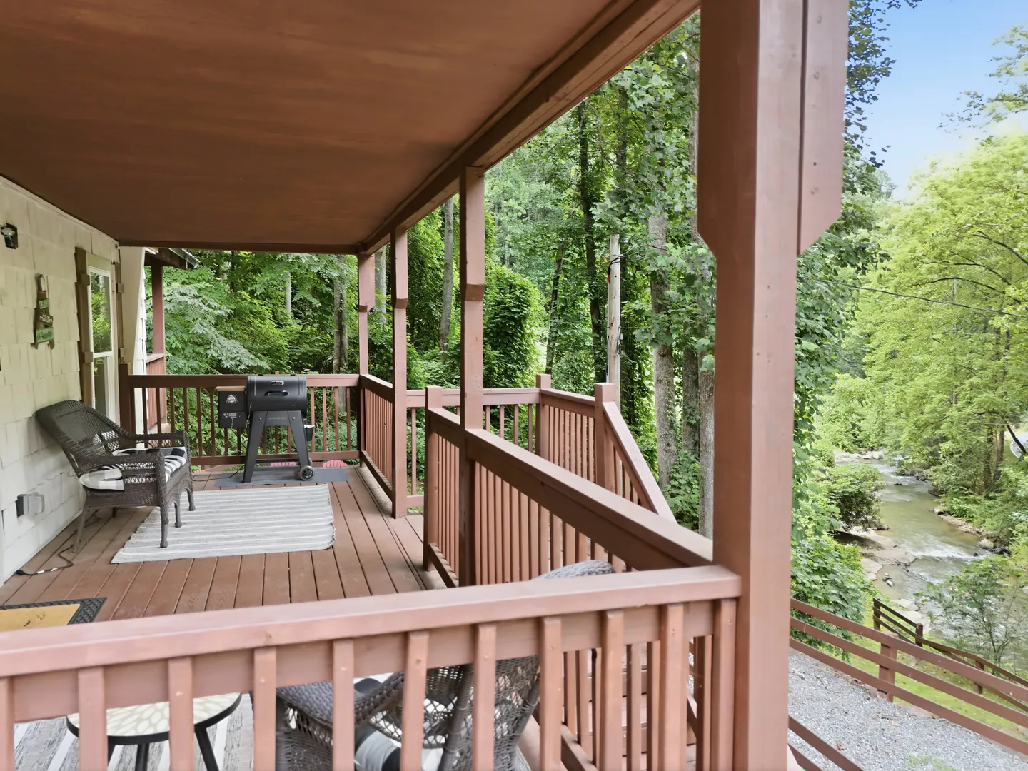 Covered porch with wooden railings, wicker chairs, a barbecue grill, and a view of a stream through lush green trees.