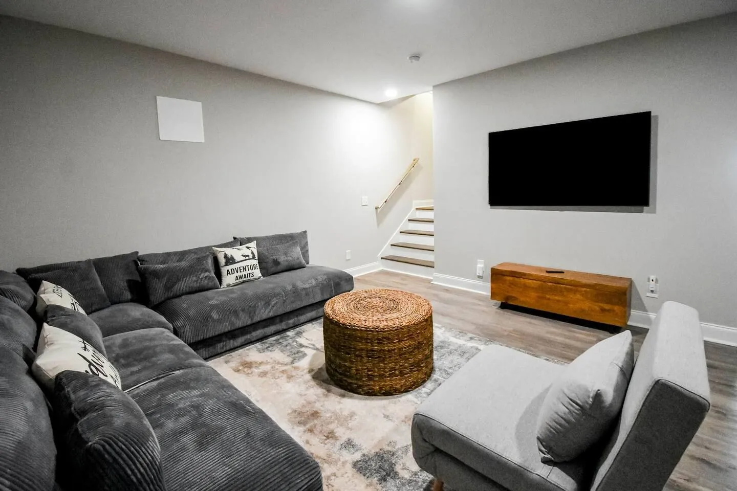 Living room featuring a large gray sectional sofa, a woven ottoman, a gray accent chair, a wall-mounted TV, and stairs leading up in the background.