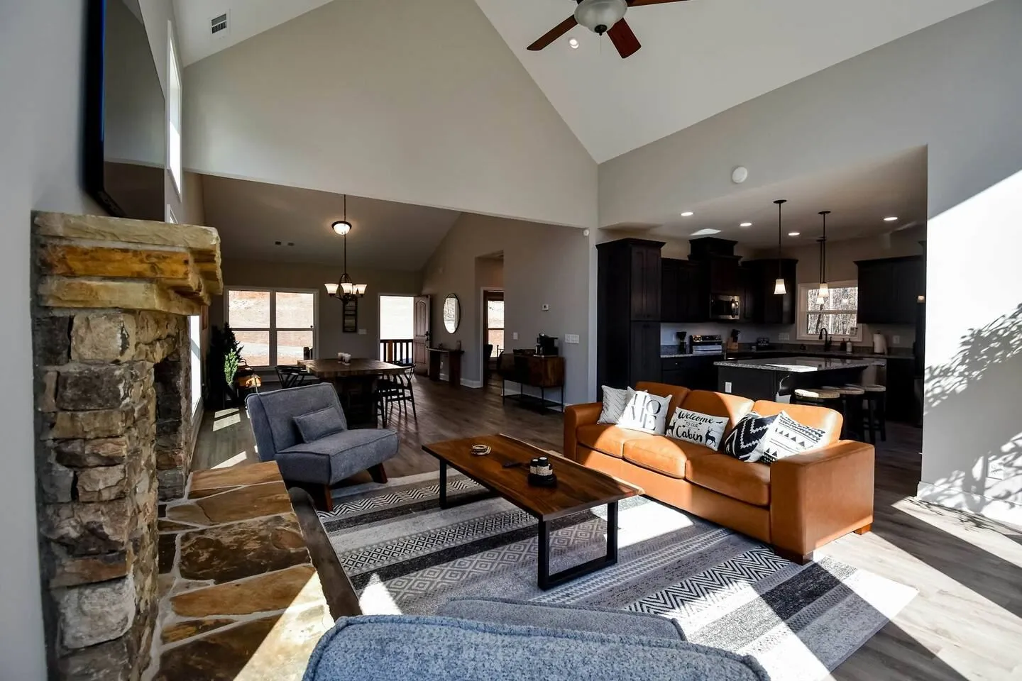 Open-concept living room with vaulted ceiling, stone fireplace, leather sofa, and views into the dining area and kitchen.