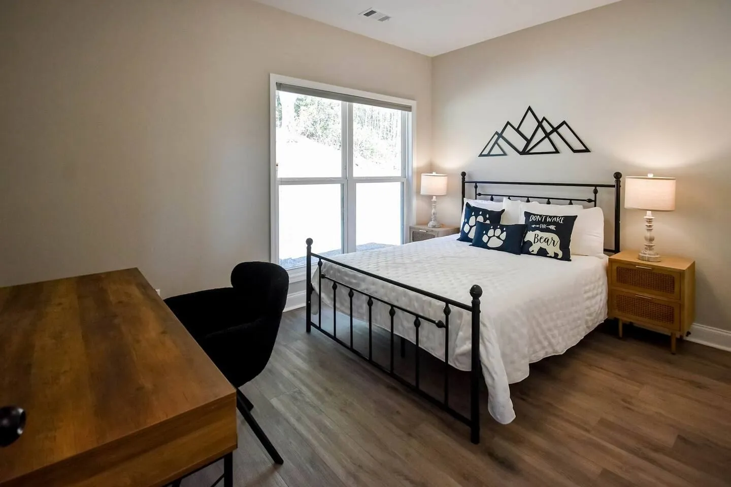Bedroom featuring a black metal bed with mountain-themed pillows, a wooden nightstand with lamp, a window, and a wooden desk with a black chair.