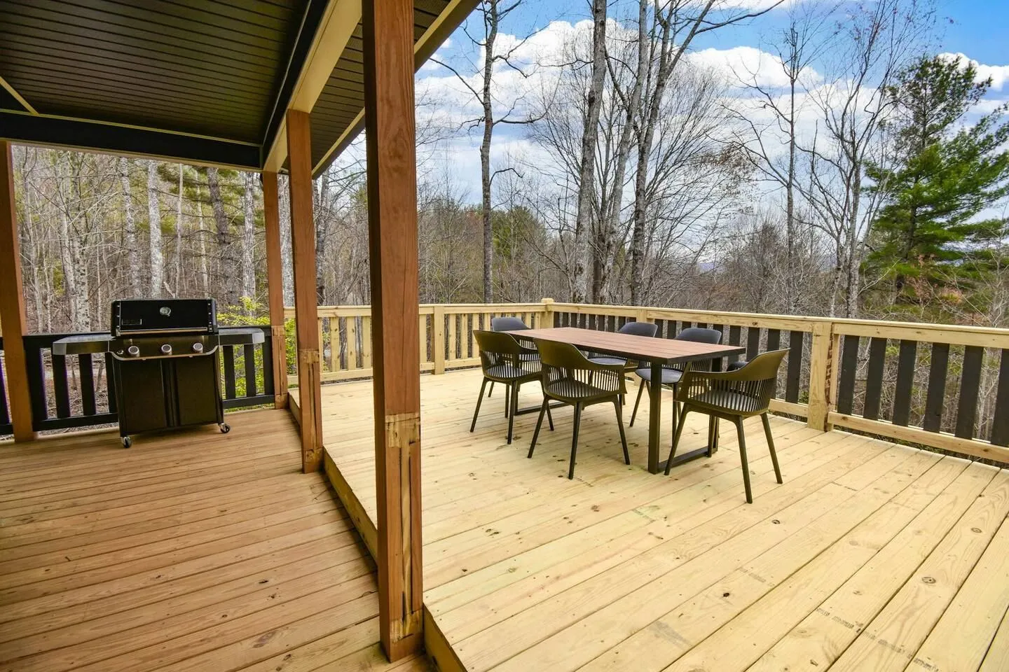 An outdoor wooden deck with a dining table, chairs, a barbecue grill, and a forest view in the background.
