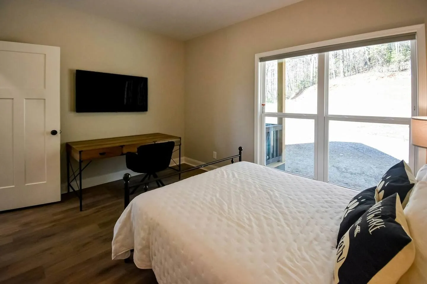 Bedroom with a bed in the foreground, a wall-mounted TV, a desk with an office chair, and a large sliding glass door looking outdoors.