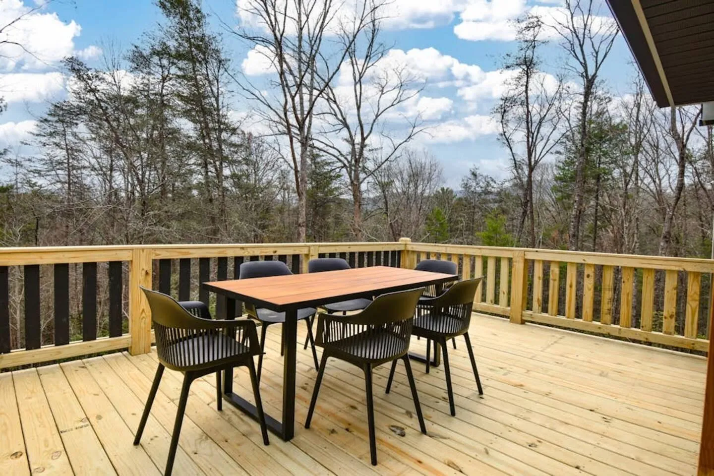 Outdoor wooden deck featuring a dining table with chairs, surrounded by railings and overlooking trees under a partly cloudy sky.