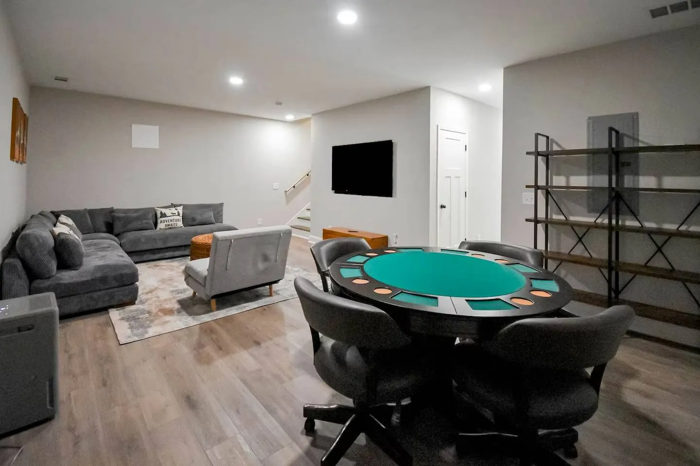 Finished basement game room featuring a round poker table with rolling chairs, a sectional sofa area with a wall-mounted TV, and light wood-look flooring.